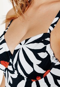 Bikini top noir et blanc à motifs avec design push-up, présentant des motifs floraux et des accents rouges. Le tissu est lisse avec des bretelles réglables.