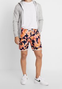 Costumi da bagno da uomo con un motivo floreale tropicale in arancione e rosa su uno sfondo blu navy, con rifiniture bianche e cintura regolabile.