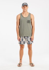 Groene tanktop met logo, gebloemde zwembroek in blauw en crème, flip-flops en een grijze pet. Model staat met de armen langs het lichaam.