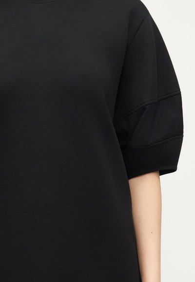 Μαύρο κοντομάνικο τοπ με λεπτομέρεια ραφής με πιέτες στο πάνω μέρος του μπράτσου, φορεμένο από άτομο με ανοιχτόχρωμο δέρμα σε απλό φόντο.