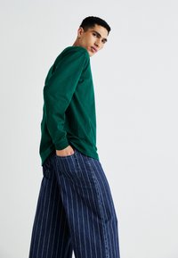 Green long-sleeve shirt paired with wide-legged, blue pinstriped pants. The shirt has a relaxed fit, while the pants feature a classic denim texture.