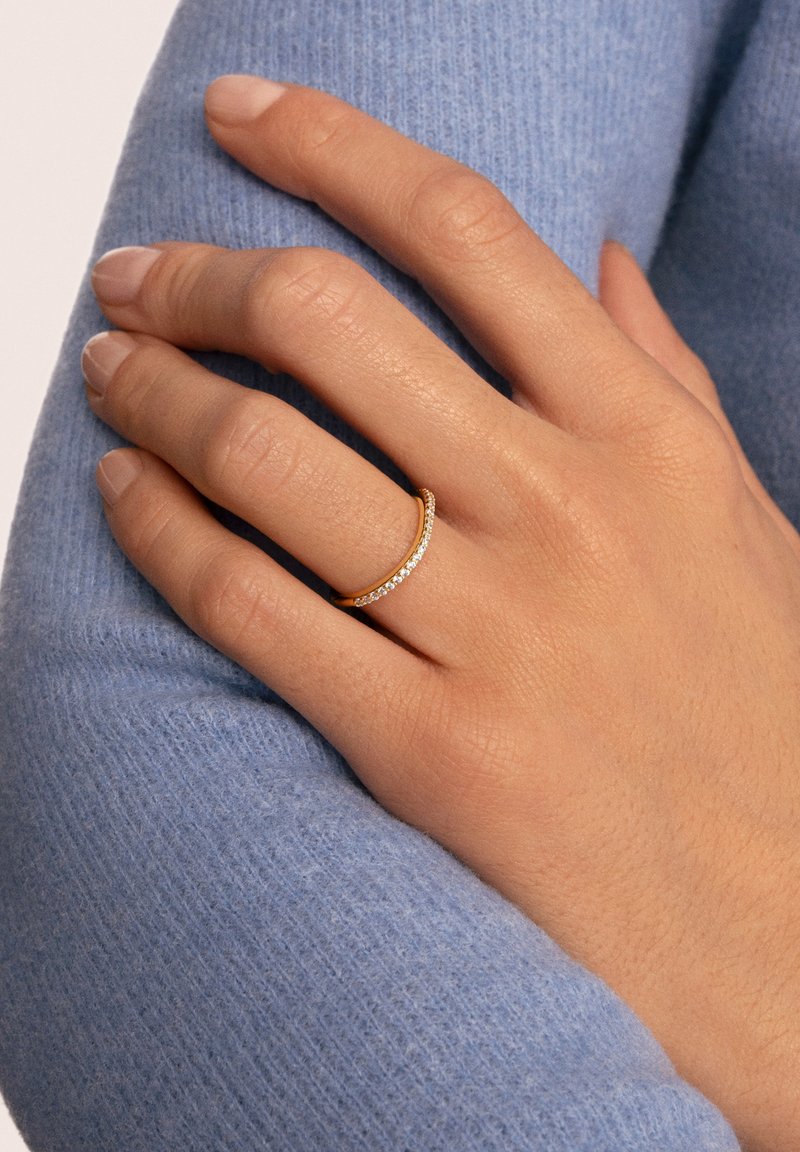 Goldring mit kleinen klaren Steinen, auf einer Hand präsentiert, die auf einem weichen, hellblauen Strickstoff ruht. Glatte und polierte Oberfläche.