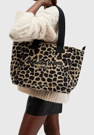 Osoba nosząca kremowy dzianinowy sweter i czarną spódnicę, trzymająca dużą torbę typu tote z nadrukiem w cętki lamparta, z czarnymi uchwytami i logo "ALLSAINTS LONDON".