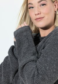 Blondýnka v stříbrných náušnicích ve tvaru kruhu a přes oversized šedém pleteném svetru, jemně držící rukáv blízko obličeje a mírně se usmívající.