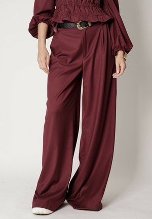 Burgundowe spodnie szerokie wykonane z gładkiego materiału; charakteryzują się wysokim stanem i luźnym krojem, podkreślone czarnym paskiem i białymi trampkami.