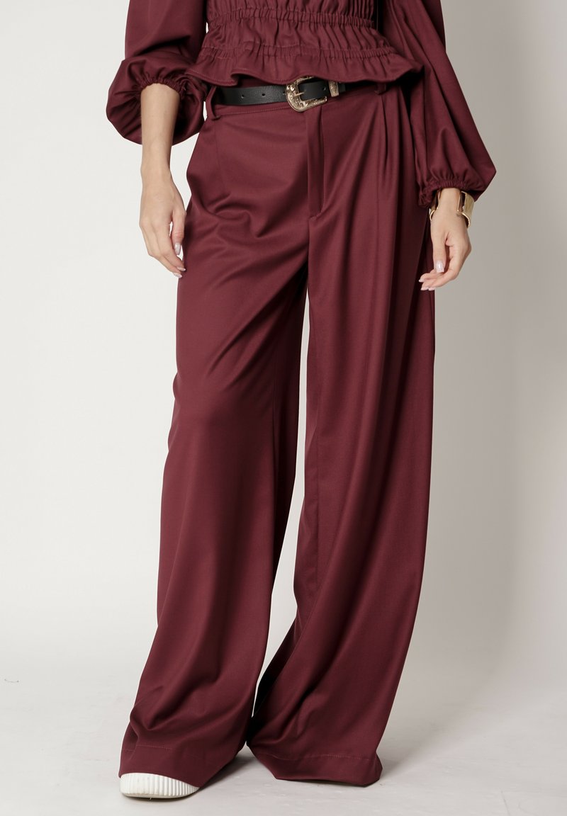 Burgundowe spodnie szerokie wykonane z gładkiego materiału; charakteryzują się wysokim stanem i luźnym krojem, podkreślone czarnym paskiem i białymi trampkami.