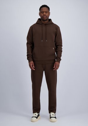 Ein Mann steht frontal, trägt einen braunen Hoodie und passende Hose mit weißen Sneakern, vor einem schlichten hellen Hintergrund.