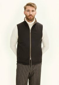 Schwarze ärmellose Weste mit einem Reißverschluss vorne, hellbeigem Kragen und zwei Seitentaschen. Getragen über einem cremefarbenen Rollkragenpullover und kombiniert mit gestreiften Hosen.