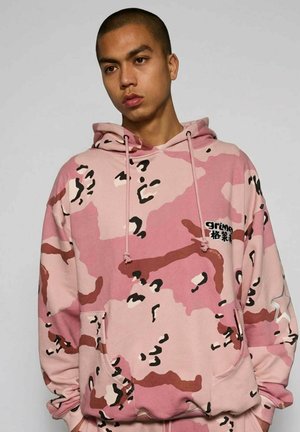 Jeune homme portant un sweat à capuche camouflage rose avec des motifs noirs et marron, une poche avant et un patch étoile sur la manche gauche.