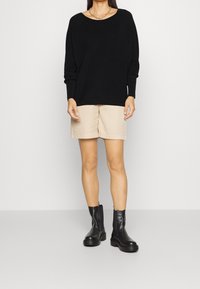 Schwarzer, übergroßer Pullover mit weitem Halsausschnitt, kombiniert mit beigen Shorts und schwarzen klobigen Stiefeletten. Glatte Textur und lässige Passform.