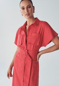 Czerwona sukienka koszulowa z krótkimi rękawami, dwoma kieszeniami na piersi, paskiem w talii oraz długości do połowy łydki. Gładka tekstura materiału.