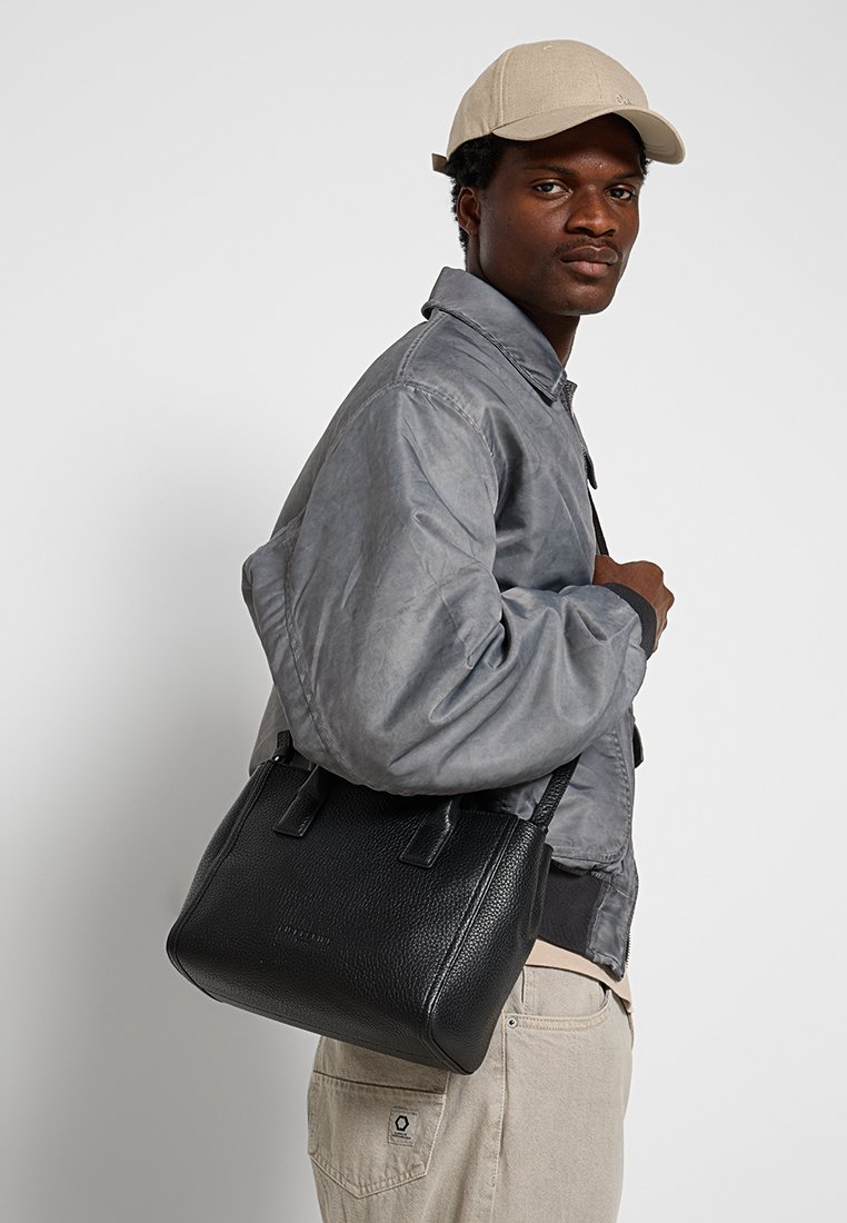 Schwarze Ledertasche mit strukturierter Oberfläche und zwei Tragegriffen oben, quer über den Körper getragen. Modell in einer grauen Jacke und beigem Hut, vor einem neutralen Hintergrund.