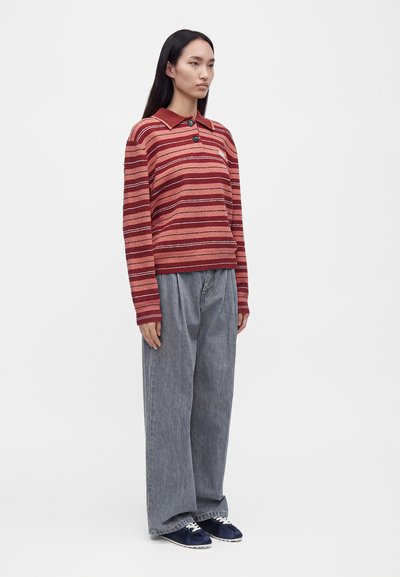 Femme aux longs cheveux foncés portant un pull rayé rouge, un pantalon gris ample et des baskets bleu marine, debout sur un fond blanc uni.