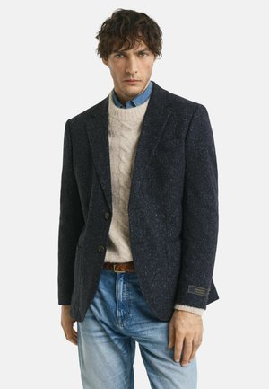 Blazer en tweed marine à deux boutons et tissu texturé, porté sur un pull en maille torsadée crème et un jean en denim. Comprend une étiquette de poignet de marque.