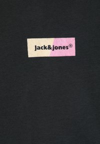 Svart t-skjorte i stoff med en rektangulær logo-patch i pastellgul og rosa med "Jack & Jones"-tekst i fet svart.