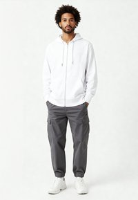 Felpa bianca con zip e coulisse, abbinata a pantaloni cargo grigi con tasche laterali, indossata con sneaker bianche.