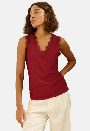 Vrouw met een mouwloze donkerrode top met kanten rand en crème kleurige broek, staand met één hand op haar heup tegen een witte achtergrond.