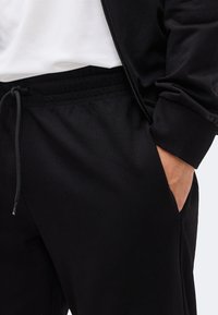 Κοντινό πλάνο ενός ατόμου που φορά μαύρο παντελόνι με κορδόνι και μαύρο μπουφάν με λευκό πουκάμισο, το χέρι ξεκουράζεται στην τσέπη του παντελονιού.