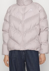 Ljusrosa vadderad jacka med rutmönster, hög krage, dragkedja och elastiska ärmslut. Har sidofickor och en logodetalj.