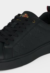 Baskets en cuir noir avec motif embossé, lacets noirs, œillet doré et logo blason coloré sur la languette.