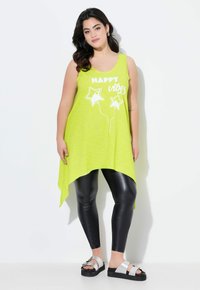 Top sin mangas de color verde lima con el texto "vibras FELICES" y gráficos de estrellas, combinado con leggings negros brillantes y sandalias de tiras plateadas; dobladillo asimétrico.