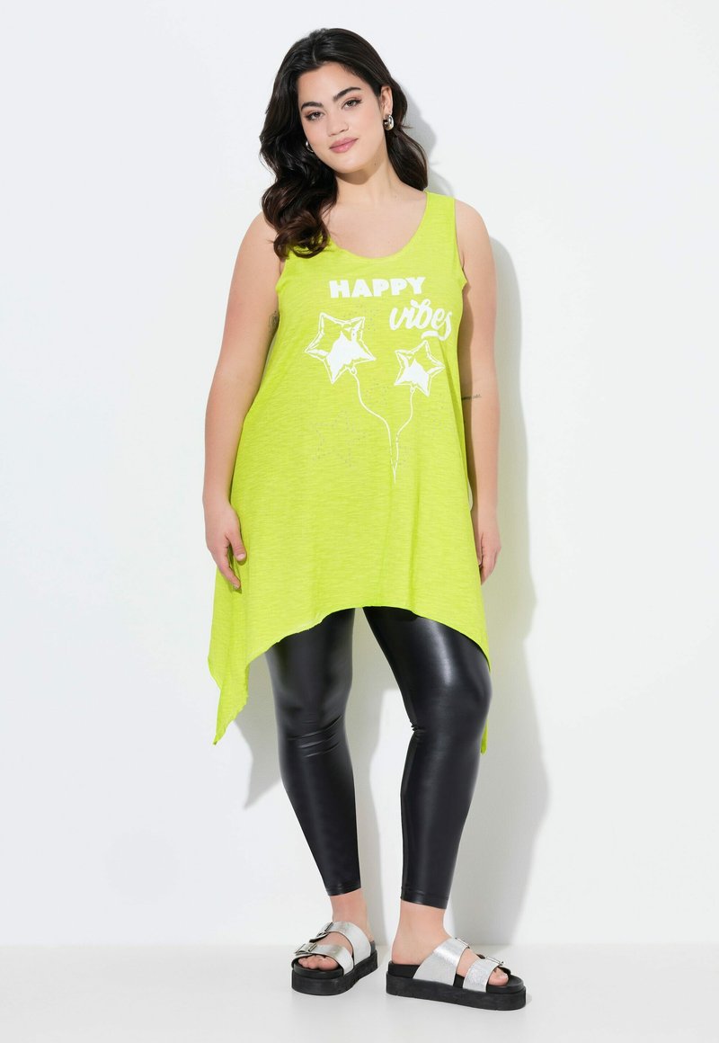 Top sin mangas de color verde lima con el texto "vibras FELICES" y gráficos de estrellas, combinado con leggings negros brillantes y sandalias de tiras plateadas; dobladillo asimétrico.