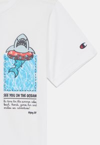 T-shirt en coton blanc avec un graphique de requin dans un flotteur rouge, fond d'eau bleue et texte noir. Logo Champion sur la manche.