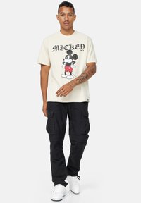 Camiseta de algodón color crema con un gráfico de Mickey Mouse en pantalones cortos rojos y zapatos negros, combinada con pantalones cargo negros y zapatillas blancas.