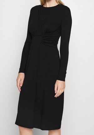 Kobieta ubrana w czarną sukienkę z długim rękawem, sięgającą do kolan, z marszczeniem w talii i okrągłym dekoltem.