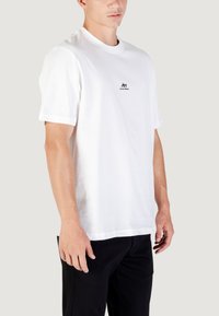 Camiseta de algodón blanca con mangas cortas, cuello redondo y logo negro en el pecho. Combinada con pantalones negros, de textura suave y ajuste casual.