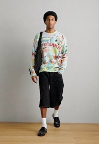 Πολύχρωμο πουλόβερ με αφηρημένα σχέδια, συνδυασμένο με μαύρα σορτς μέχρι το γόνατο, λευκές κάλτσες και μαύρα παπούτσια χωρίς κορδόνια. Φορώντας μαύρη τσάντα.