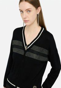 Cardigan nero con scollo a V, dettagli a righe bianche e chiusura con zip. Decorato con strass sulla parte superiore anteriore.