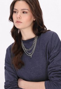 Trójwarstwowy srebrny naszyjnik łańcuszkowy z mieszanymi ogniwami, noszony na ciemnogranatowym swetrze. Gładka faktura, z detalem w postaci kokardki i małymi zawieszkami.