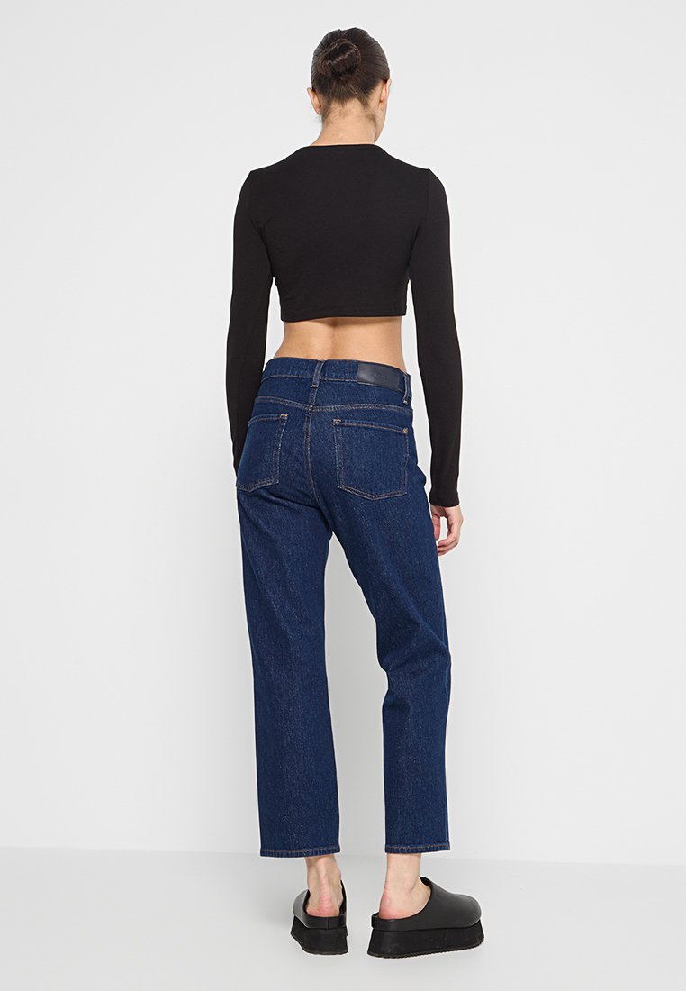 Un crop top noir à manches longues, associé à un jean en denim bleu foncé taille haute. Le jean a une coupe décontractée et des poches arrière, avec un accent en cuir.