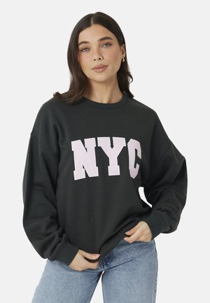Kobieta o brązowych włosach, ubrana w czarną bluzę z "NYC" dużymi białymi literami i jasnoniebieskie jeansy, stojąca na jednolitym tle.