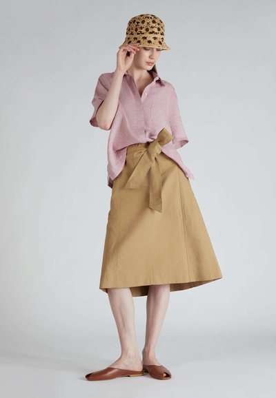 Camicia di lino rosa chiaro con taschino, abbinata a una gonna A-line color khaki con un nastro in vita. Scarpe aperte con punta scoperta di colore marrone.