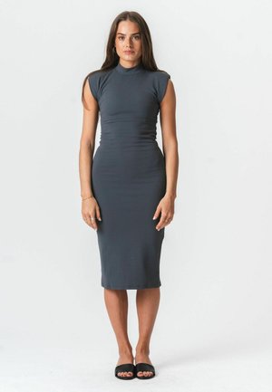 Nő áll, sötétszürke, testhezálló, ujjatlan midi ruhát és fekete, nyitott orrú papucsot visel, sima fehér háttér előtt.