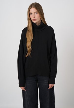 Jonge vrouw met lang blond haar, dragend een losse zwarte coltrui en donkere wijde broek, staand tegen een effen achtergrond.