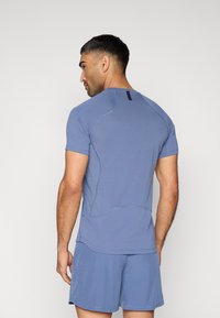 Αθλητικό κοντομάνικο μπλουζάκι από μαλακό, υφασμάτινο μπλε ύφασμα με λεπτομέρειες ραφών και μικρό μαύρο διακοσμητικό στο πίσω μέρος του λαιμού.