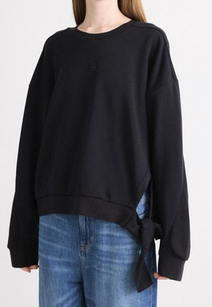 Sweat-shirt noir oversize à manches longues avec détail de nouage sur le côté, porté avec un jean bleu par une personne aux cheveux longs.