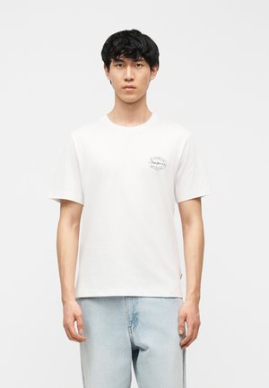 Młody mężczyzna o krótkich czarnych włosach, ubrany w gładki biały t-shirt z małym logo na lewej piersi oraz jasnoniebieskie dżinsy, stoi na jednolitym tle.