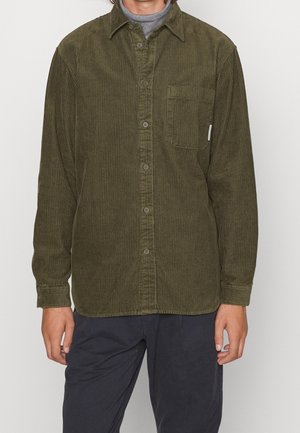 Mann trägt ein olivgrünes Cordhemd über einem grauen Rollkragenpullover, kombiniert mit dunkler Hose, steht vor einfachem Hintergrund.