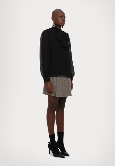 Schwarze, durchsichtige Bluse mit einer Schleifenbindung, kombiniert mit einem grauen Karominirock. Schwarze Ankle Boots mit hohem Absatz und Riemenakzent.