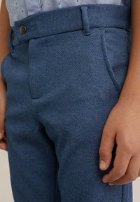 Blauwe textuurbroek met een gladde afwerking, voorzakken en een knoopsluiting in de taille, met een close-up van de stof en hand.