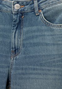 Prednji krupni plan plavih traperica koji prikazuje pojas, metalni gumb, petlju za remen, patentni zatvarač i detalje prednjeg džepa.