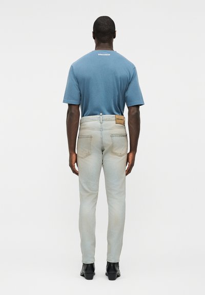 Homme portant un jean gris clair délavé et un t-shirt bleu Dsquared2, debout de dos sur un fond blanc uni.