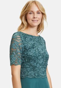 Tealowy top z koronką o wzorze kwiatowym, z falbaniastymi rękawami, nakładany na jednolitą, tealową tkaninę. Posiada okrągły dekolt i delikatną strukturę.