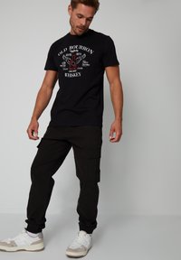 Zwarte katoenen T-shirt met een whiskey-graphic en tekst, gecombineerd met zwarte cargobroeken met meerdere zakken, en witte sneakers met beige accenten.