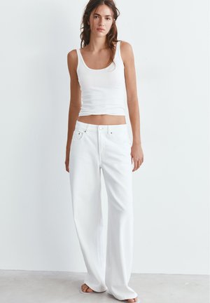 Jonge vrouw staand met een mouwloze witte tanktop en losse witte broek tegen een effen lichte achtergrond.