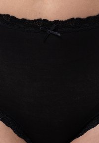 Slip nera a vita alta con rifiniture in pizzo e dettaglio di piccolo fiocco in vita, indossata sulla parte inferiore del corpo di una persona.